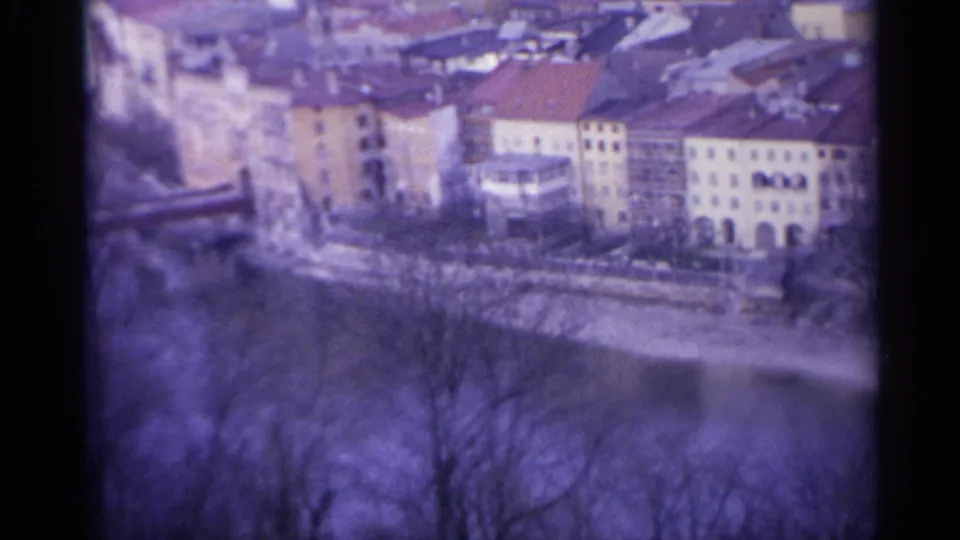 1961 8mm frame from Spain with riverside buildings, red roofs, arched lower floors, bare branches, and soft film grain.