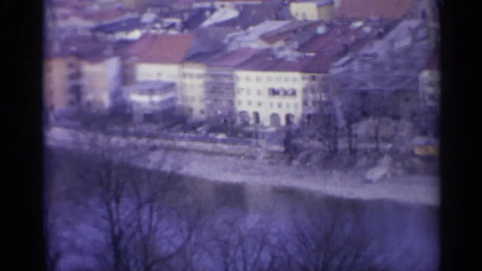 1961 Spain 8mm frame showing old riverside buildings with red-tiled roofs behind bare trees and water below.