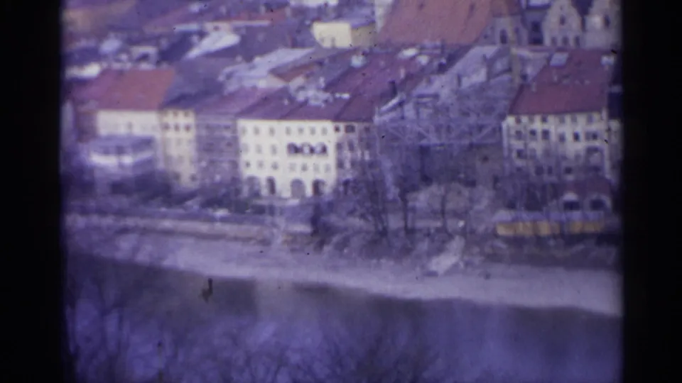 1961 Spain home-movie frame with pale town buildings, red roofs, leafless trees and a riverbank below.