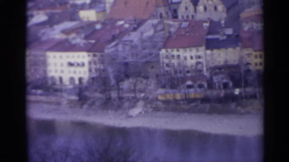 1961 Spain 8mm view of pale riverside buildings, red roofs, bare trees and a yellow vehicle near the bank.