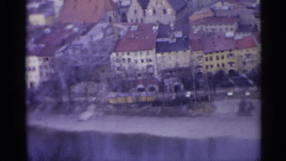 1961 Spain home-movie frame showing a wider riverside town section with old buildings, bare trees and a yellow vehicle.