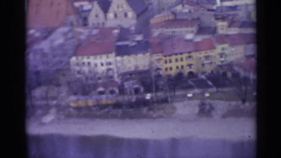 1961 8mm view of a Spanish town with pastel facades, red-tiled roofs, bare trees, parked cars, and a water edge in the foreground.