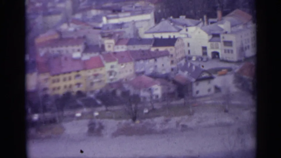 Final 8mm frame of a Spanish town in 1961, with pale buildings, red roofs, bare trees, quiet roadway, and soft color fading.