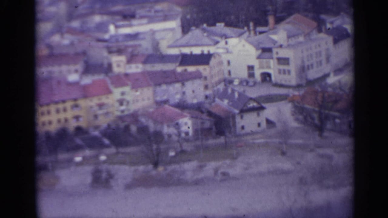 1961 Spain 8mm frame showing an elevated view over clustered town buildings, red tile roofs, open ground, and bare trees near hills.