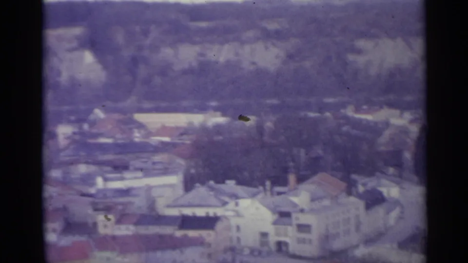 Soft, color-shifted 8mm frame of a Spanish village edge with roofs, trees, and a pale rocky hillside beyond.