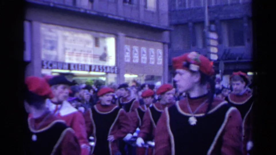 1961 Spain street parade frame with marchers in red hats and dark outfits passing an urban storefront crowd.