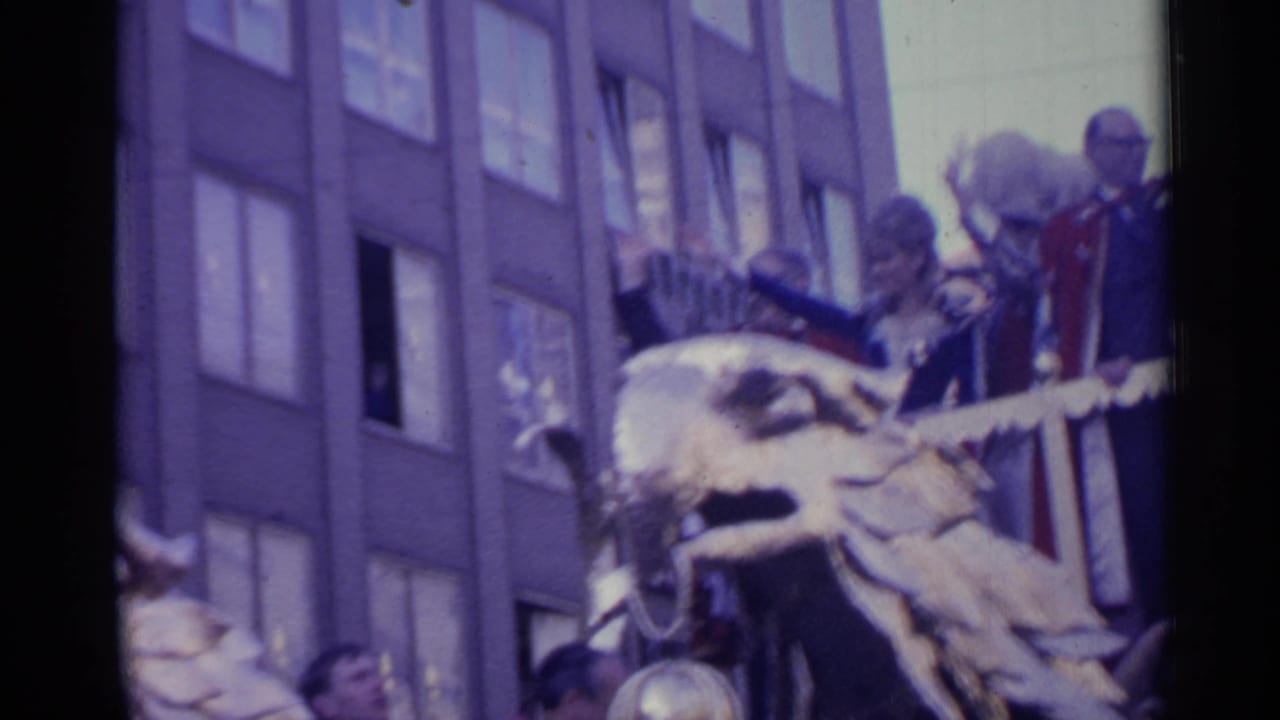 Soft 8mm frame of a decorated parade float in Spain, with an eagle ornament, formal riders, a building facade, and partial crowd faces.
