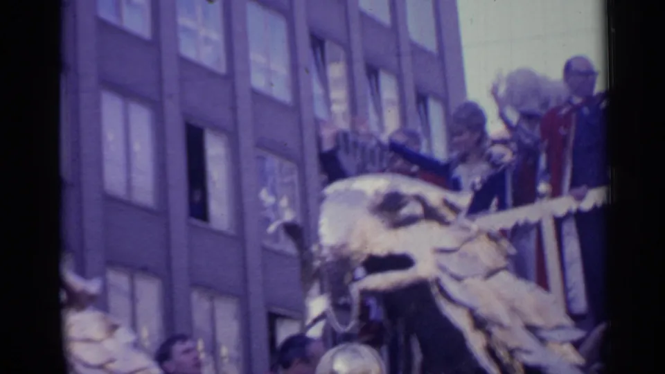 Soft 8mm frame of a decorated parade float in Spain, with an eagle ornament, formal riders, a building facade, and partial crowd faces.