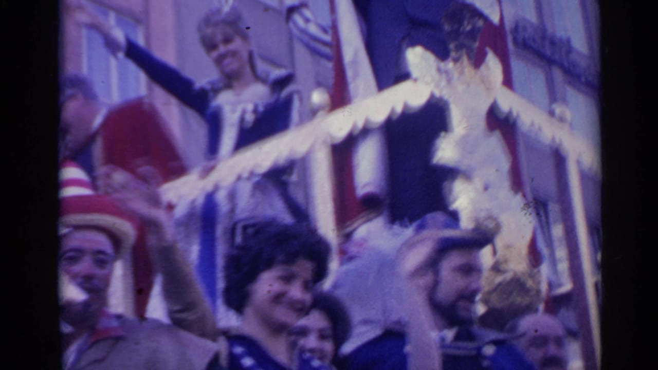 Costumed adults on the Spanish parade float smile and wave, including a woman in dark blue and a man with a beard near the rail.