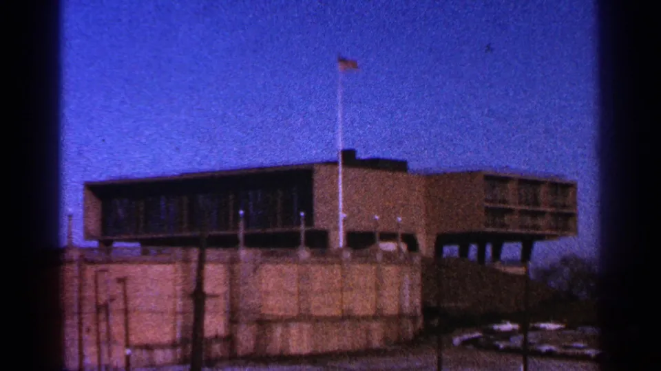 1964 Milwaukee frame with a tall flagpole in front of a brown building, broad dark windows, concrete base, and blue sky.