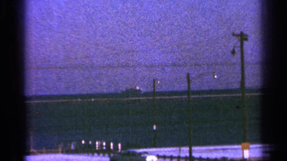 Milwaukee-associated 1964 home-movie frame with a distant ship on the horizon and poles along the waterfront foreground.