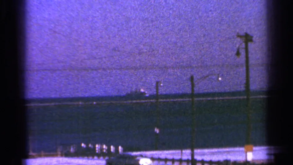 1964 Milwaukee-associated 8mm frame showing a distant ship across the water with foreground poles and visible film texture.