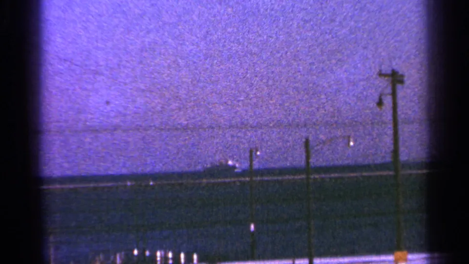 Milwaukee-associated 1964 shoreline frame with a distant ship, utility poles, dark water, and pronounced 8mm frame edges.