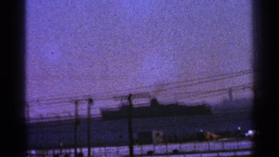 Grainy 8mm frame of a large ship at the Milwaukee port, with foreground utility lines, poles, and a small waterfront structure.