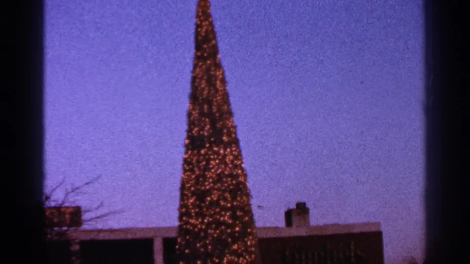 1964 Milwaukee home-movie frame showing the lit Christmas tree above low buildings and bare branches at dusk.