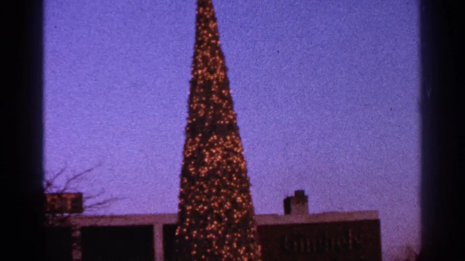1964 Milwaukee home-movie frame showing a tall lit Christmas tree rising above a department store exterior at twilight.
