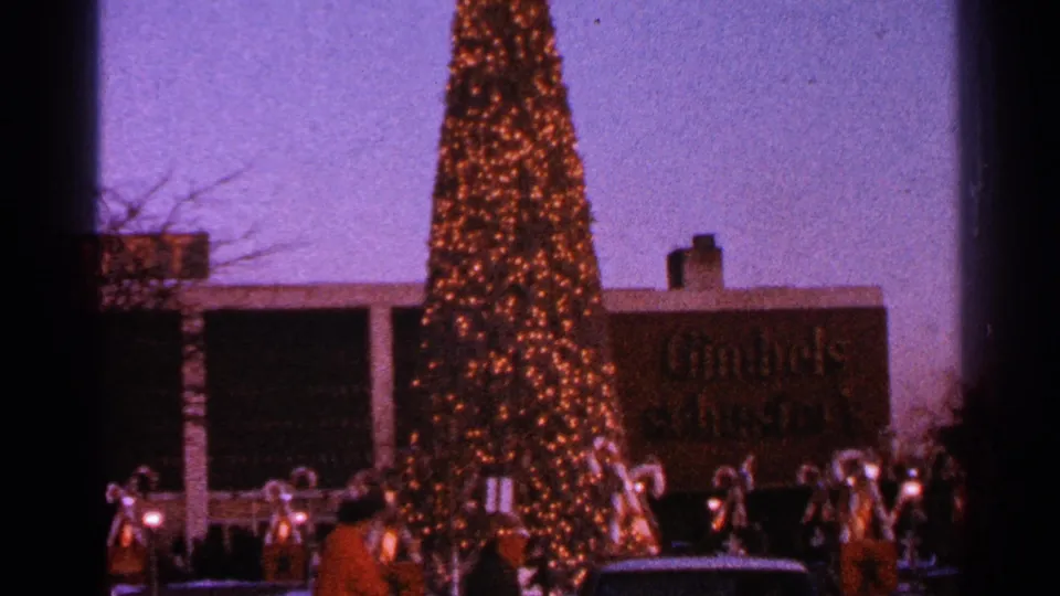 1964 Milwaukee home-movie frame showing the lower portion of a towering lit tree and holiday figures in front of a Gimbels storefront.