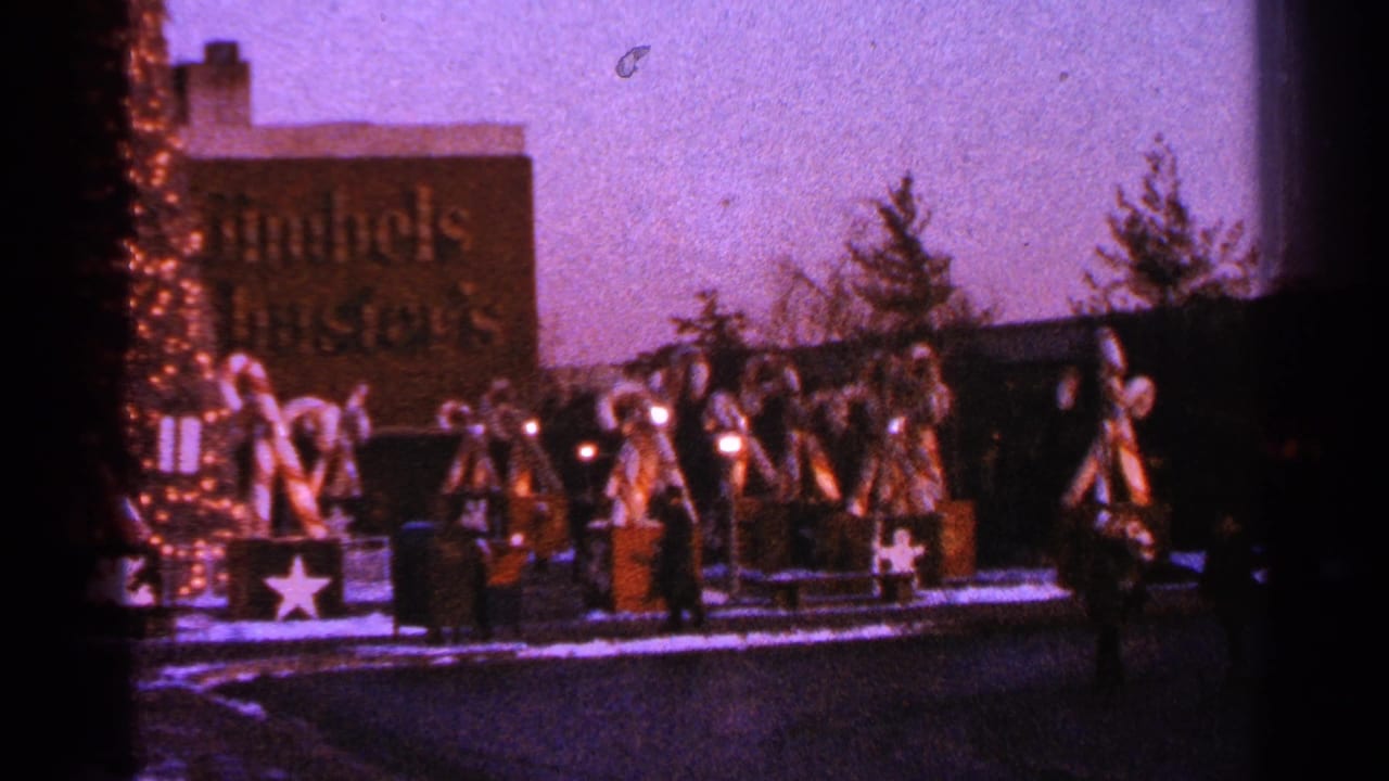 1964 Milwaukee home-movie frame showing a snowy holiday display with large candy cane decorations, bows, lights, pedestrians, and a visible sign.