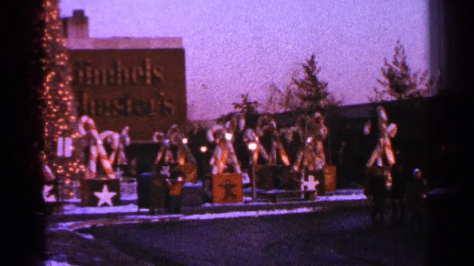 1964 Milwaukee frame of a public Christmas display with oversized candy cane figures, lighted trim, snow, pedestrians, and a commercial sign behind it.
