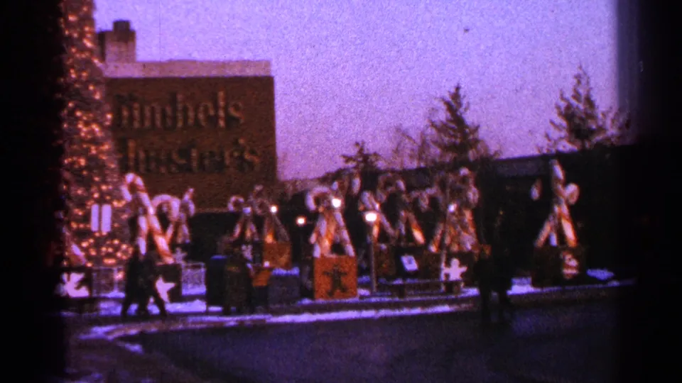 Wide 1964 Milwaukee home-movie view of a snowy candy cane holiday display with bows, lights, pedestrians, and a partially readable commercial sign.