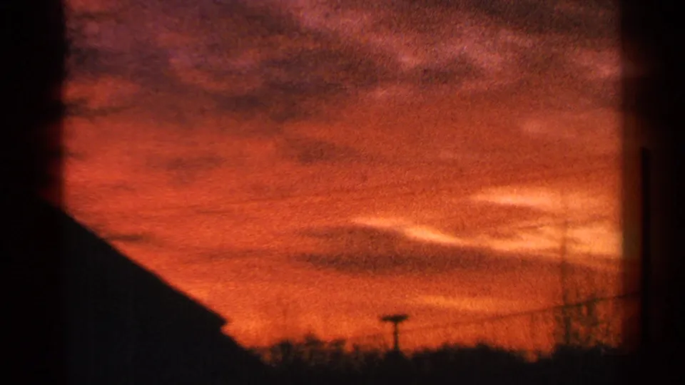 1964 Milwaukee 8mm frame of orange-red evening clouds over a dark horizon with a sloped roofline, wires, and bare trees.
