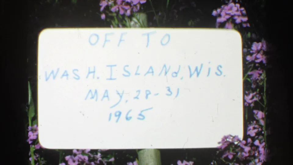 Mid-1960s home-movie frame showing a white handwritten sign for Washington Island, Wisconsin, with flowers around the edges.