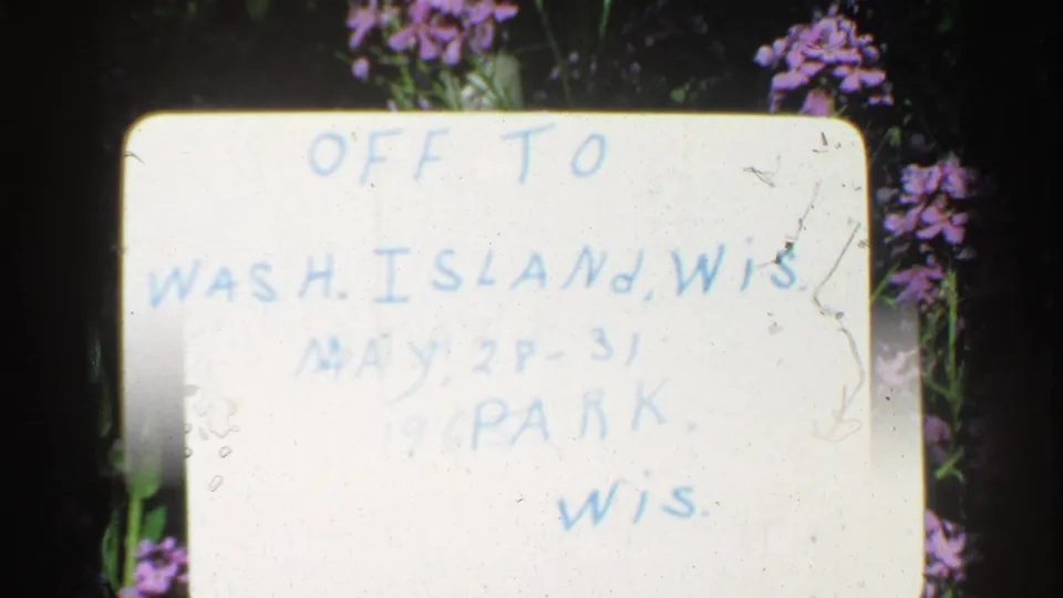 8mm frame of the white Washington Island, Wisconsin trip sign, partly obscured by exposure and scratches, with pink flowers nearby.