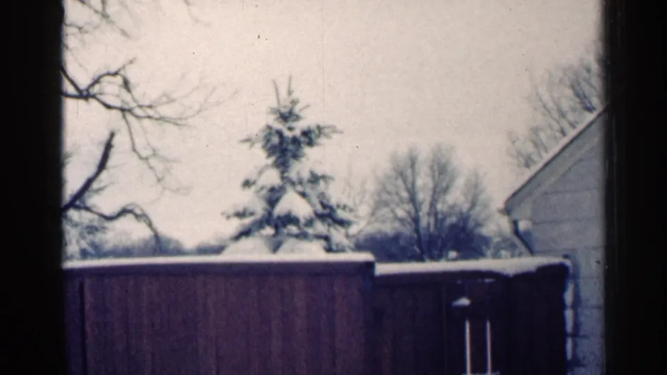 1964 Minnesota 8mm frame showing a wooden fence in a snowy yard with a small evergreen, bare trees, and part of a house at right.