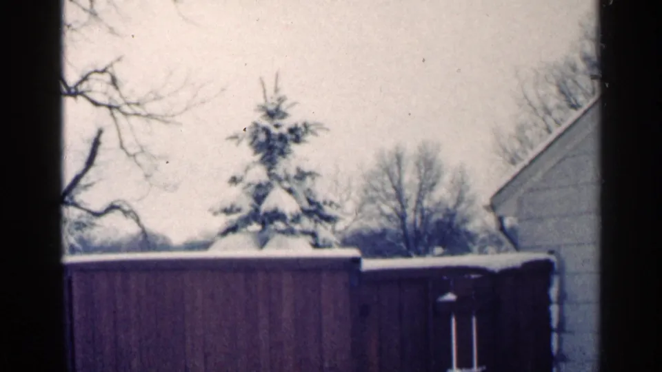 1964 Minnesota 8mm frame looking across a snow-covered fenced yard toward bare trees, a small evergreen, and a house edge.