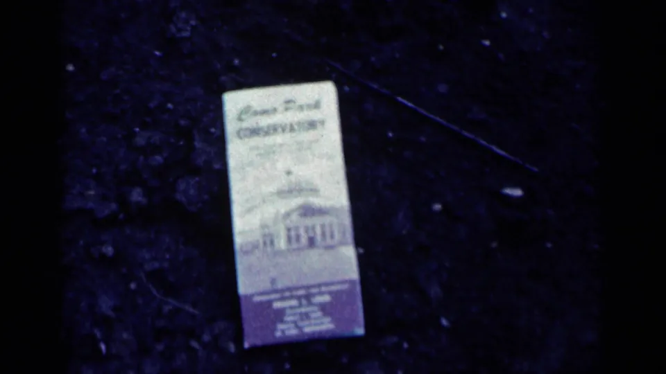1964 Minnesota home-movie frame showing a Como Park Conservatory matchbook on dark ground, with printed text visible but partly soft.