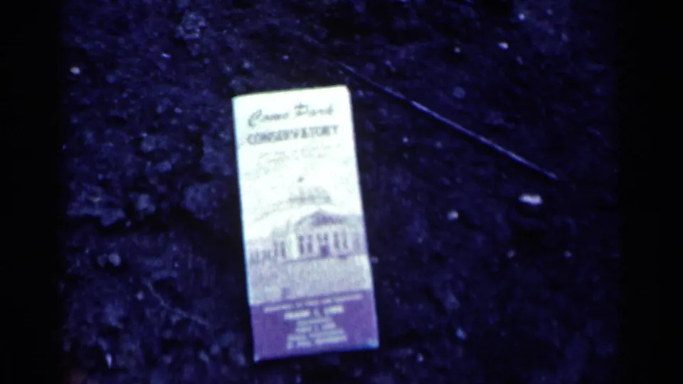 1964 Minnesota 8mm frame showing a Como Park Conservatory brochure on dark pavement with soft focus and blue color shift.
