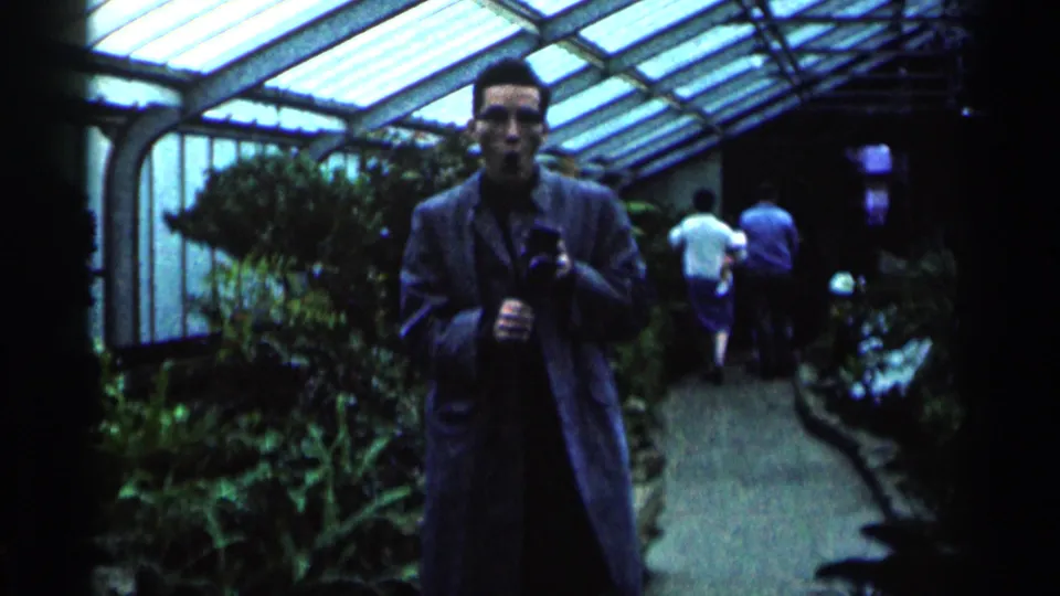 A man wearing glasses and a long coat faces the camera in a glass-roof greenhouse, with visitors walking along the path behind him.