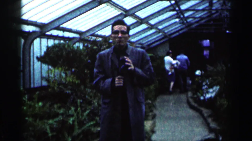 The man remains centered on the greenhouse walkway, holding a camera as visitors move in the background beneath the glass roof.