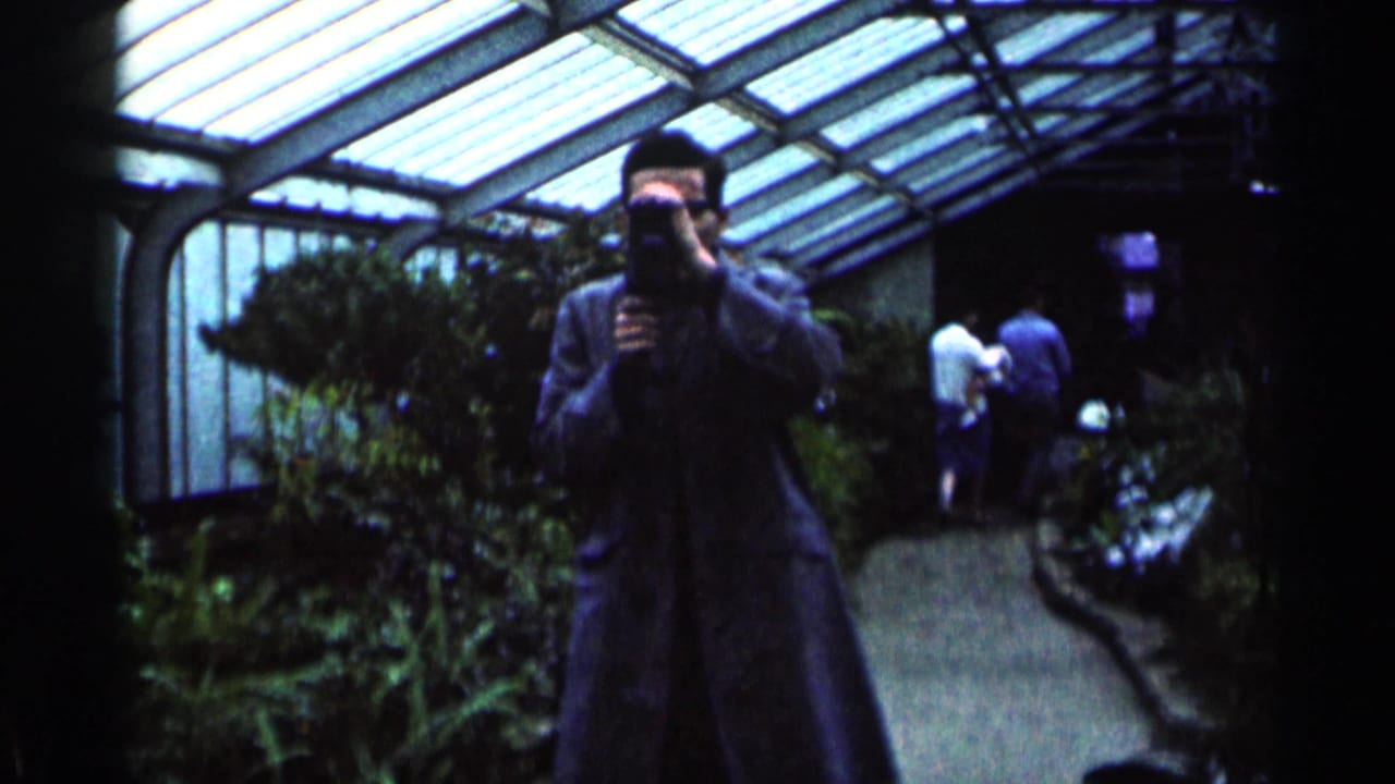 1964 Minnesota 8mm frame showing a man holding a camera in a greenhouse walkway beneath a glass roof, with foliage and visitors behind him.