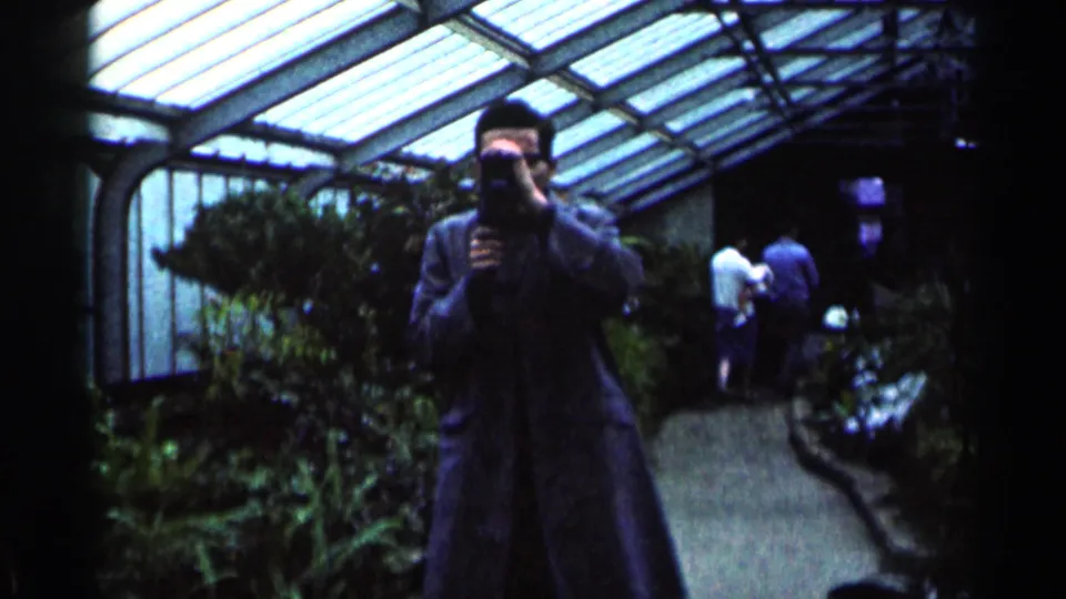 1964 Minnesota 8mm frame showing a man holding a camera in a greenhouse walkway beneath a glass roof, with foliage and visitors behind him.