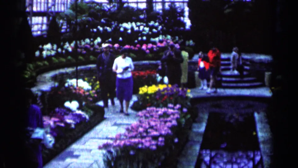 Minnesota 1964 8mm frame showing visitors beside flower beds, stone paths, and a dark sunken water channel inside a conservatory-like space.