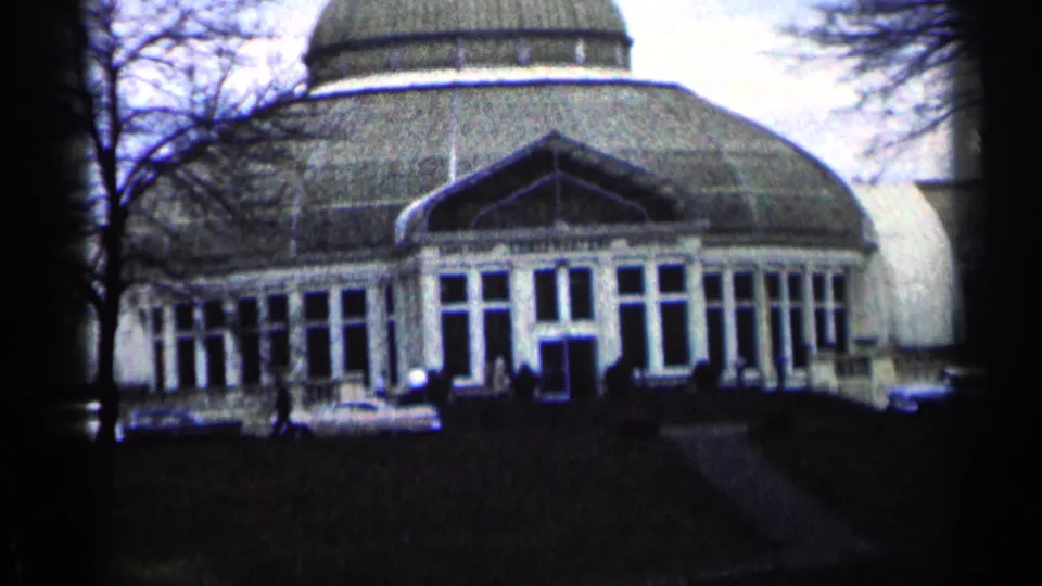 Close 1964 Minnesota home-movie frame of the domed building entrance, with white cars and small visitors below the glass facade.