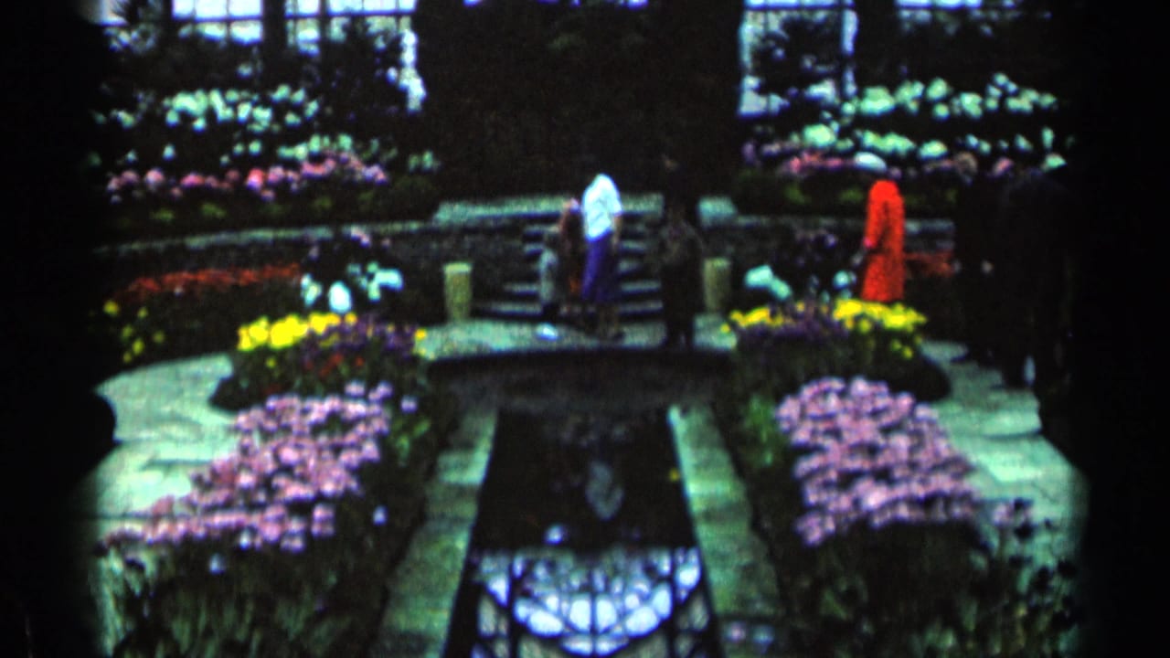 Visitors walk through a conservatory garden with colorful flower beds and a narrow pool.