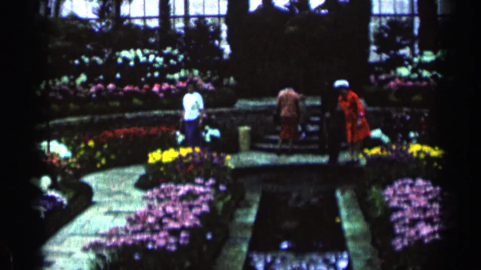 A woman in a red coat and other visitors walk through the Minnesota conservatory past dense flower beds and stone edging.