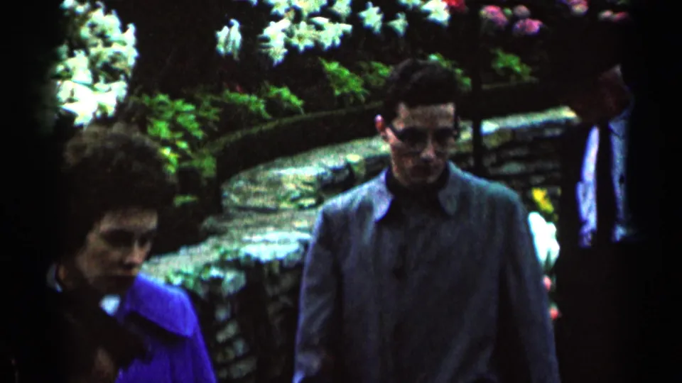1964 Minnesota 8mm frame showing adults walking through a garden with white flowers, foliage, and stone edging.