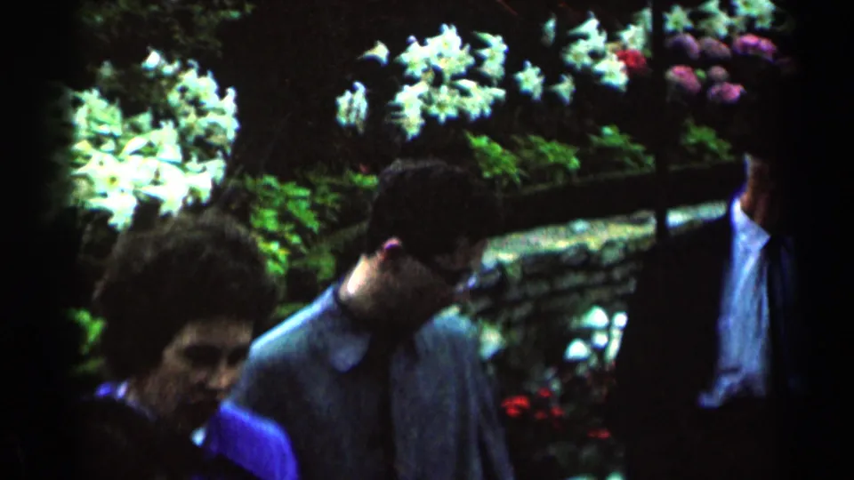 1964 Minnesota home-movie frame of adults walking through a lush garden, partly obscured by low light and film softness.