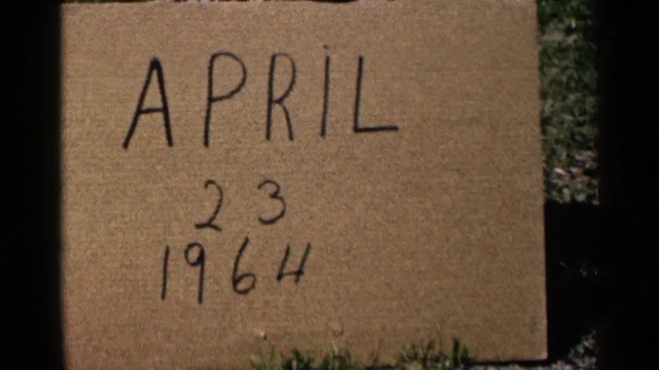 Close 8mm frame of a handwritten April 23, 1964 cardboard sign with grass visible around the edges.