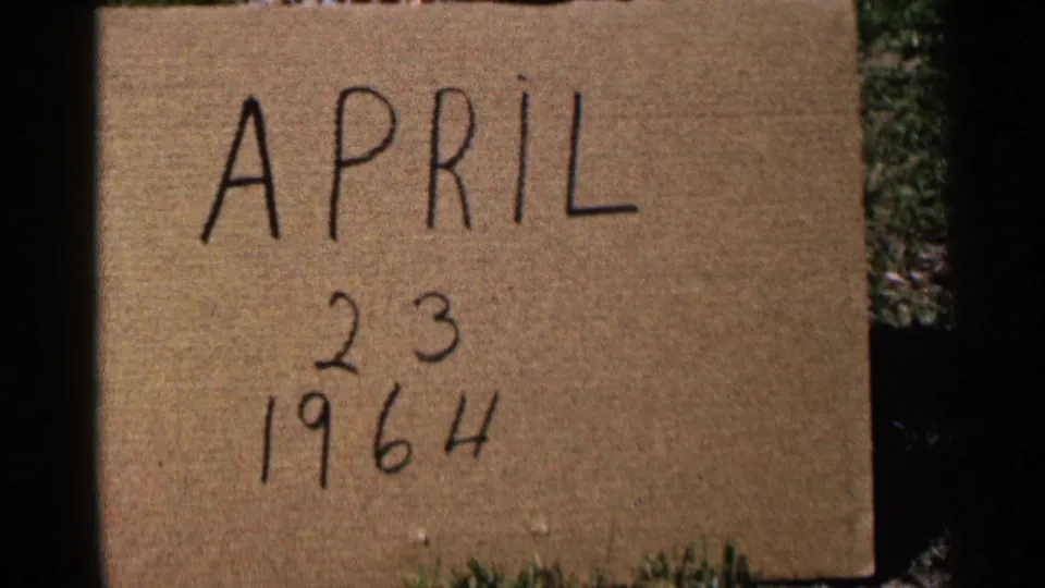 Outdoor 8mm frame of the April 23, 1964 cardboard date card, with grass and shadow at the frame edge.