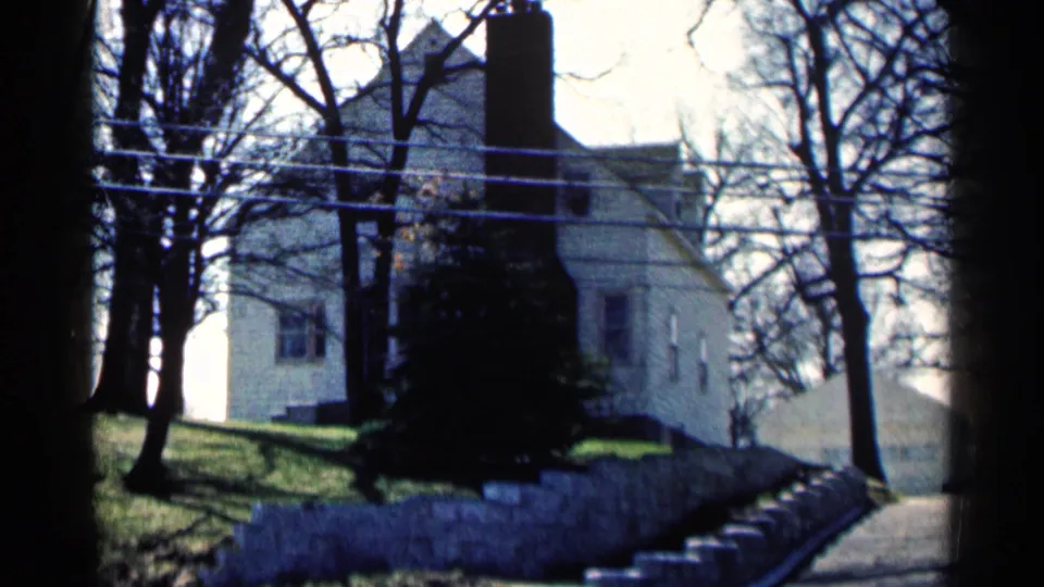 1964 Minnesota home-movie view of a white two-story house behind winter trees, with a chimney and stone wall.