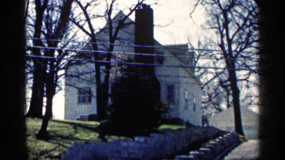 Soft 8mm frame of the Minnesota house exterior, showing clapboard siding, chimney, trees, power lines, and wall.