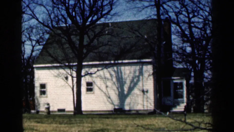 Rural Minnesota house exterior in 1964, with bare branches in the foreground and a dark roof above white siding.