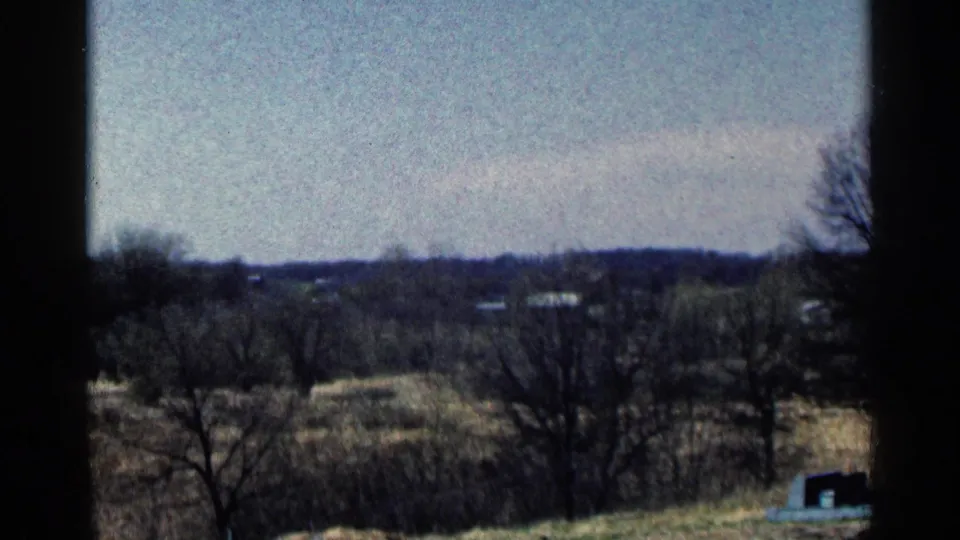 1964 Minnesota countryside view with open fields, leafless trees, low hills, and scattered distant buildings under a pale sky.