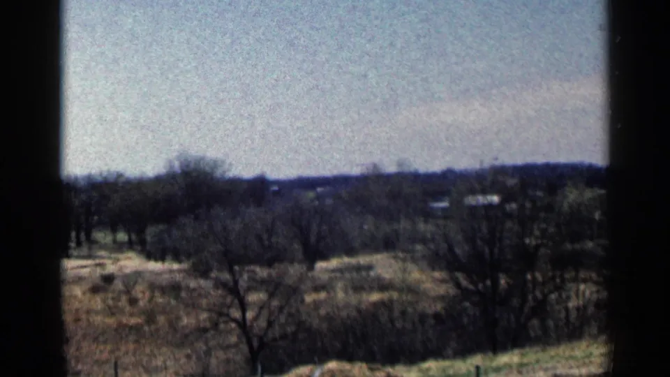 Vintage 8mm view across rural Minnesota fields, with leafless trees, distant homes, and low wooded hills in the background.