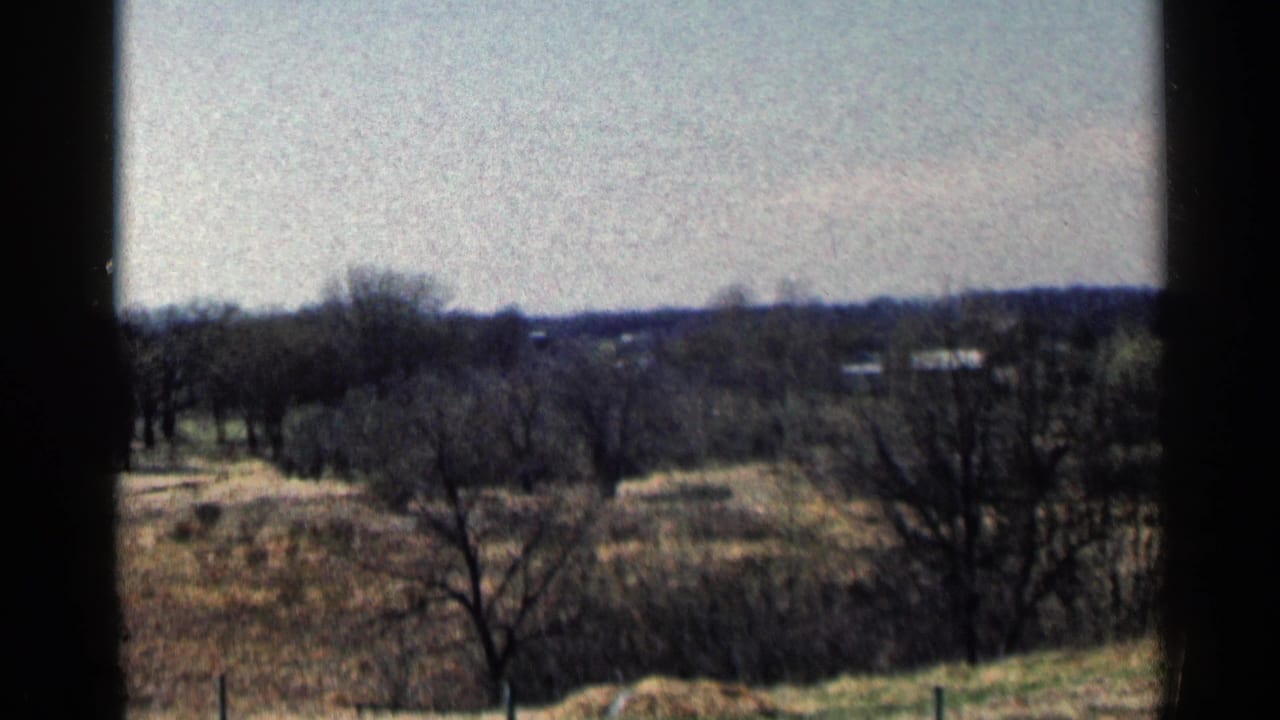 Open rural view with trees, dry ground, and low buildings near the horizon.