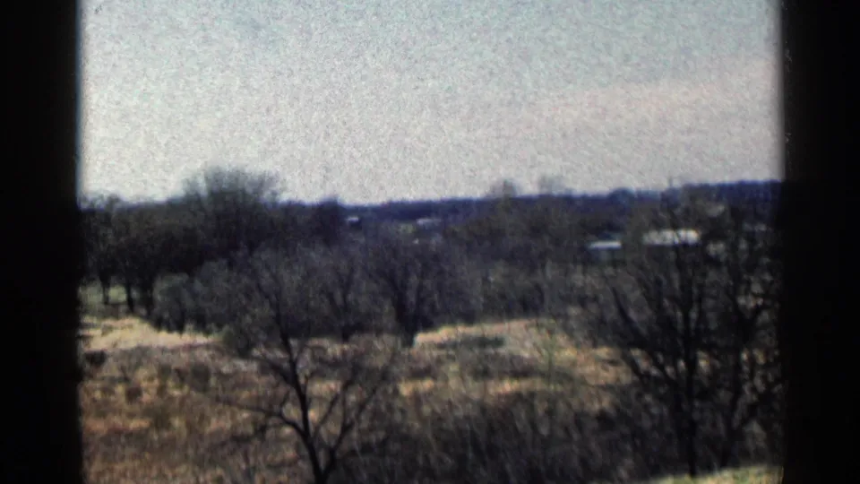 1964 Minnesota 8mm frame showing a slightly shifted view of leafless foreground trees, open dry fields, distant structures, and dark framing edges.
